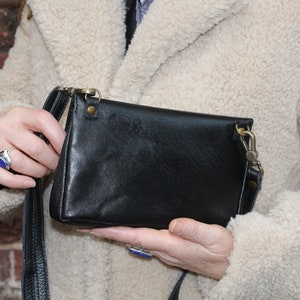 K&ouml;nnte beinhalten: Eine schwarze Leder-Umh&auml;ngetasche mit einem langen Riemen. Die Tasche hat einen Klappverschluss und einen silbernen Verschluss.