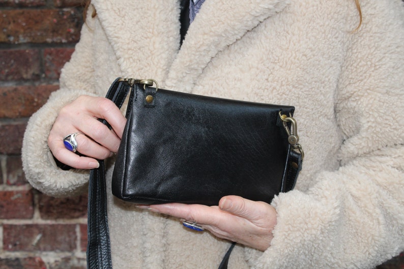 K&ouml;nnte beinhalten: Eine schwarze Leder-Umh&auml;ngetasche mit einem langen Riemen. Die Tasche hat einen Klappverschluss und einen silbernen Verschluss.