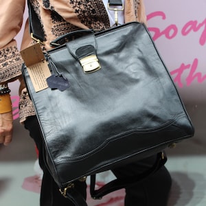 May include: A black leather satchel bag with a gold-coloured clasp and hardware. The bag has a structured design with a top handle and a shoulder strap. A tag is attached to the bag. The background is a pink colour with the words "Soap Leather".