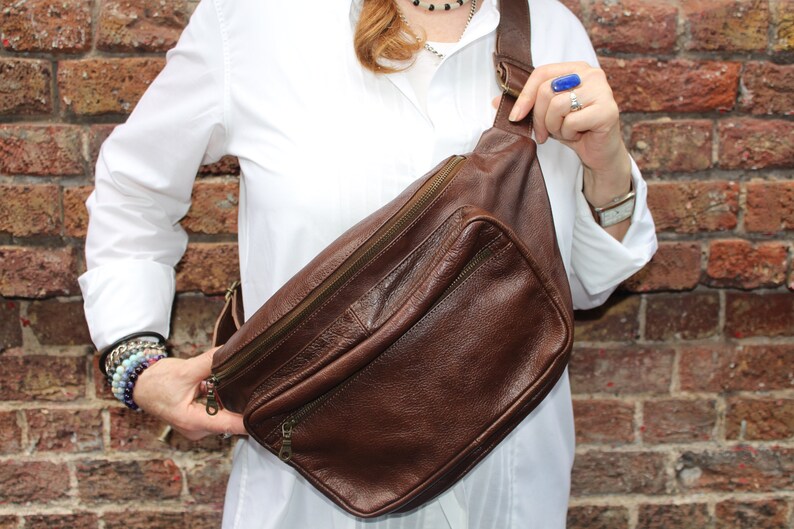 K&ouml;nnte beinhalten: Eine braune Leder-Bauchtasche mit einem Rei&szlig;verschlussfach und einem Riemen. Die Tasche wird &uuml;ber der Schulter getragen.