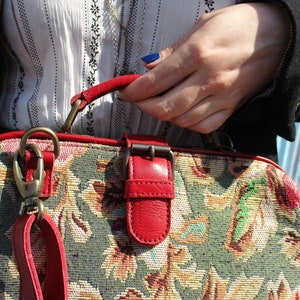 May include: A vintage-style tapestry handbag with a red leather strap, buckle, and trim. The bag has a floral pattern in shades of green, yellow, and brown.