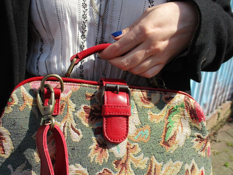 May include: A vintage-style tapestry handbag with a red leather strap, buckle, and trim. The bag has a floral pattern in shades of green, yellow, and brown.