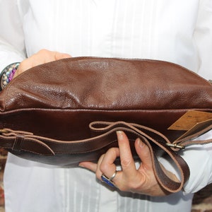 K&ouml;nnte beinhalten: Eine braune Leder-Umh&auml;ngetasche mit einem langen, verstellbaren Riemen. Die Tasche hat einen Klappenverschluss mit einer Schnalle und eine Rei&szlig;verschlusstasche auf der Vorderseite.