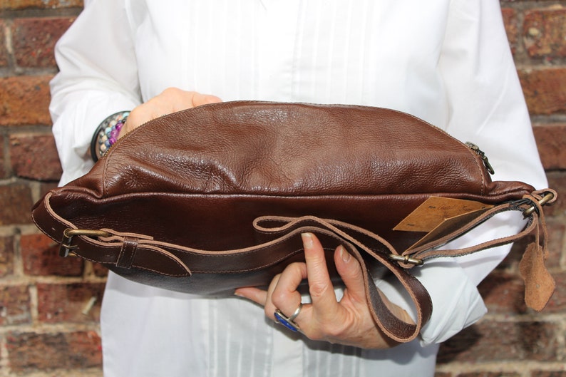 K&ouml;nnte beinhalten: Eine braune Leder-Umh&auml;ngetasche mit einem langen, verstellbaren Riemen. Die Tasche hat einen Klappenverschluss mit einer Schnalle und eine Rei&szlig;verschlusstasche auf der Vorderseite.