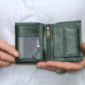May include: A dark green bi-fold wallet featuring a clear plastic ID window on the left side, a central vertical zippered compartment with a metal pull, and a small flap-covered pocket on the right side, all constructed with visible stitching.