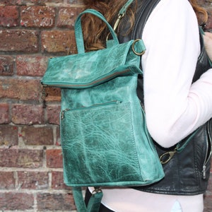 May include: A teal green leather backpack with a front zipped pocket and a top flap. The bag has a handle and adjustable straps. The backpack is approximately 30 cm wide and 36 cm tall.