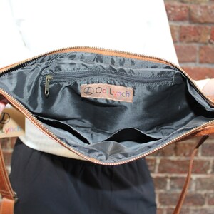 May include: A brown leather crossbody bag with a zipper closure and a black fabric interior. The bag has a small zippered pocket and a larger open pocket inside. The bag has a label that says "Odd Lynch".