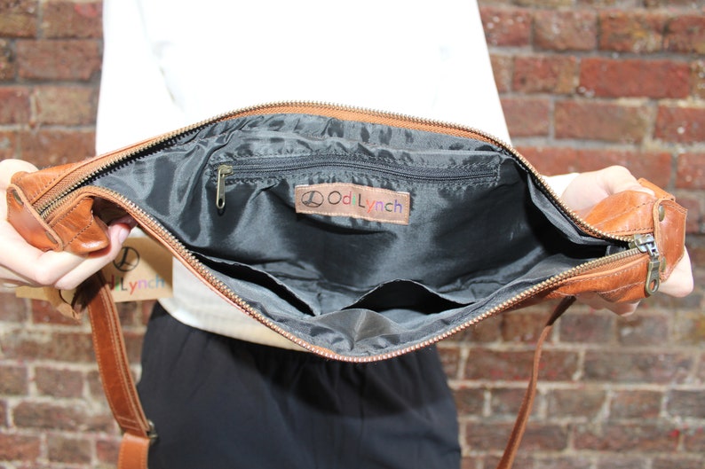 May include: A brown leather crossbody bag with a zipper closure and a black fabric interior. The bag has a small zippered pocket and a larger open pocket inside. The bag has a label that says "Odd Lynch".