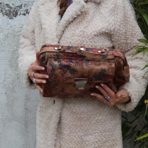 May include: A vintage handbag featuring a floral design in brown, red, and cream. The rectangular bag has a front pocket, a silver clasp, and a top handle. The bag is held against a cream-coloured coat. The bag's dimensions are approximately 30 cm in length.