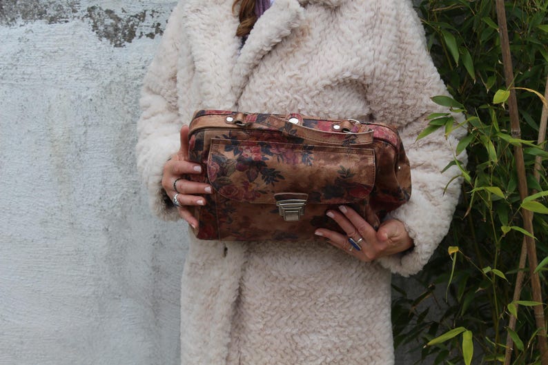 May include: A vintage handbag featuring a floral design in brown, red, and cream. The rectangular bag has a front pocket, a silver clasp, and a top handle. The bag is held against a cream-coloured coat. The bag's dimensions are approximately 30 cm in length.