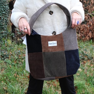 Puede incluir: Un bolso tote de pana con patchwork en tonos marrones, negros y grises. El bolso tiene una correa ancha de pana a juego y una etiqueta que dice "TinkerCreek".