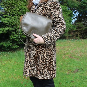 May include: A brown leather satchel bag with a flap closure and a long strap. The bag is being held by a person wearing a leopard print coat.
