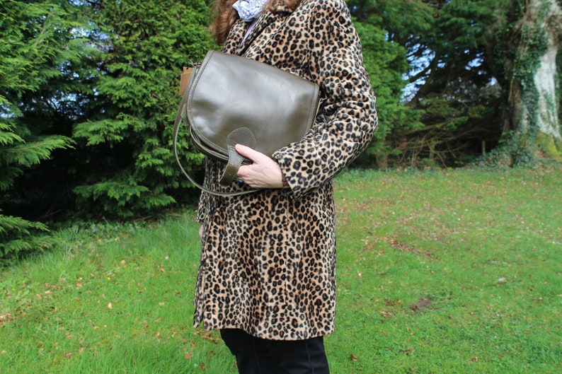 May include: A brown leather satchel bag with a flap closure and a long strap. The bag is being held by a person wearing a leopard print coat.
