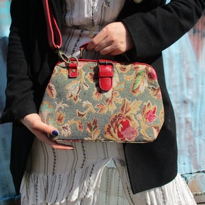 May include: A floral patterned handbag with a red leather strap and buckle closure. The bag has a vintage style and is made of tapestry fabric.