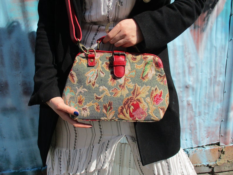 May include: A floral patterned handbag with a red leather strap and buckle closure. The bag has a vintage style and is made of tapestry fabric.
