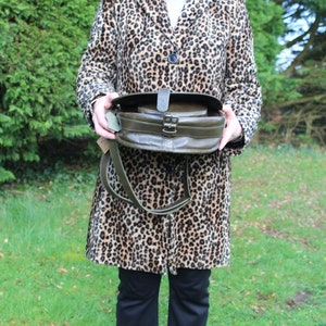 May include: A green leather satchel bag with a flap closure and a long strap. The bag is held in front of a person wearing a leopard print coat.