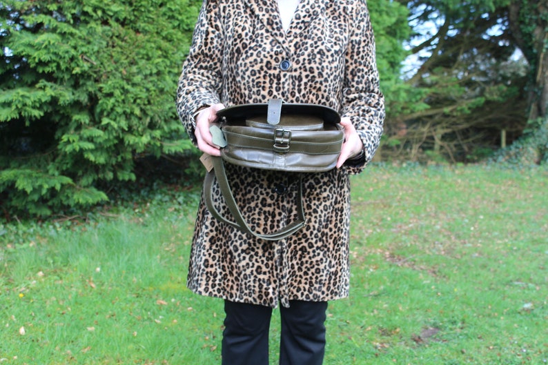 May include: A green leather satchel bag with a flap closure and a long strap. The bag is held in front of a person wearing a leopard print coat.