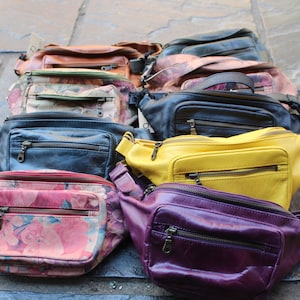 May include: A collection of ten fanny packs in various colors and patterns, including floral, leather, and solid colors. The fanny packs are arranged in a row on a gray tiled surface.