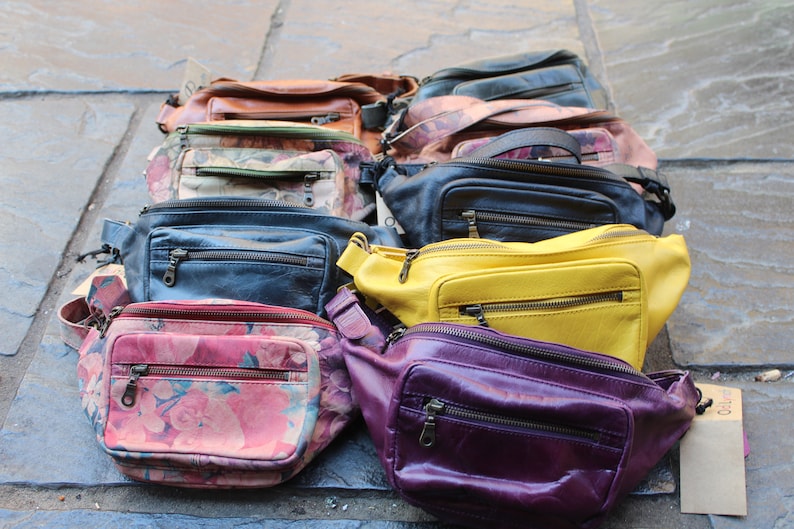 May include: A collection of ten fanny packs in various colors and patterns, including floral, leather, and solid colors. The fanny packs are arranged in a row on a gray tiled surface.