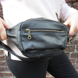 May include: A black leather fanny pack with a zipper closure and a metal zipper pull. The fanny pack is worn around the waist.