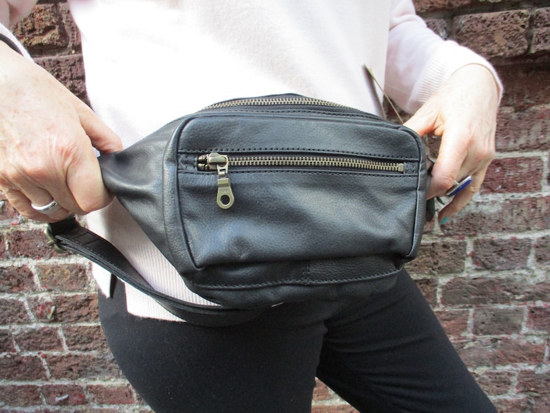 May include: A black leather fanny pack with a zipper closure and a metal zipper pull. The fanny pack is worn around the waist.