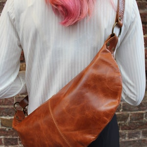 May include: A brown leather crossbody bag with a curved shape. The bag is worn by a person with pink hair.