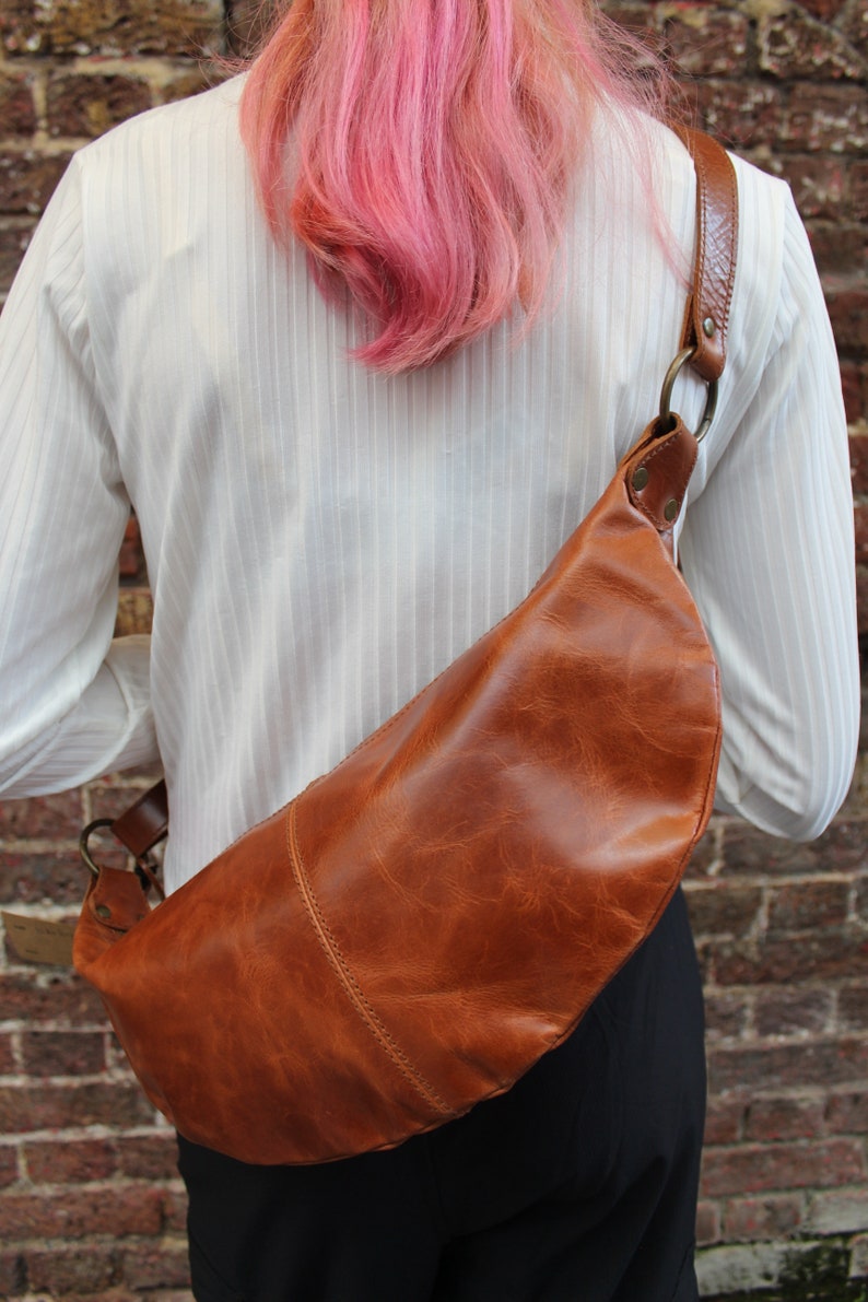 May include: A brown leather crossbody bag with a curved shape. The bag is worn by a person with pink hair.