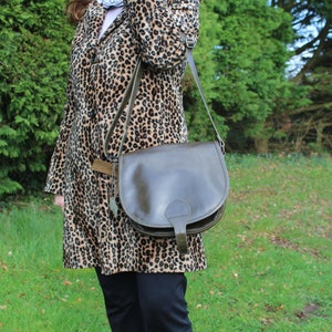 May include: A brown leather satchel bag with a flap closure and a strap. The bag is worn over the shoulder of a person wearing a leopard print coat.