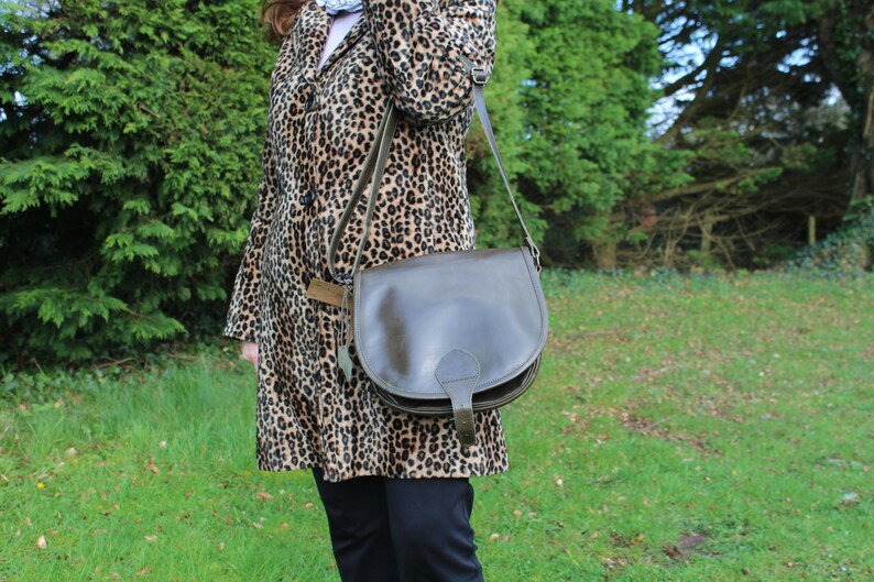 May include: A brown leather satchel bag with a flap closure and a strap. The bag is worn over the shoulder of a person wearing a leopard print coat.
