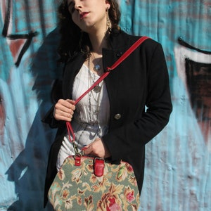 May include: A woman wearing a black coat and a white dress with a floral pattern holds a vintage-style handbag with a red strap. The handbag is made of a tapestry fabric with a floral design in shades of green, brown, and pink.