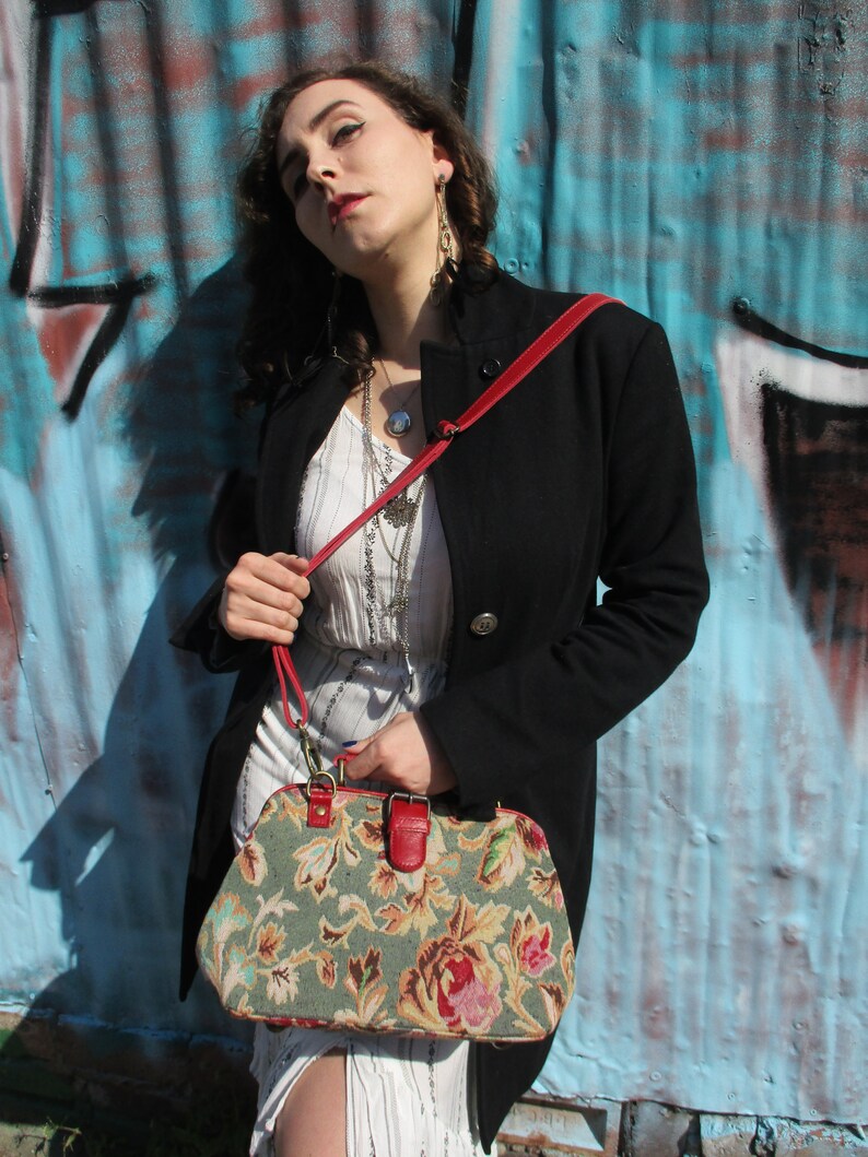 May include: A woman wearing a black coat and a white dress with a floral pattern holds a vintage-style handbag with a red strap. The handbag is made of a tapestry fabric with a floral design in shades of green, brown, and pink.