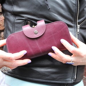 May include: A burgundy leather wallet with a flap closure and a silver button. The wallet is being held in someone's hands.