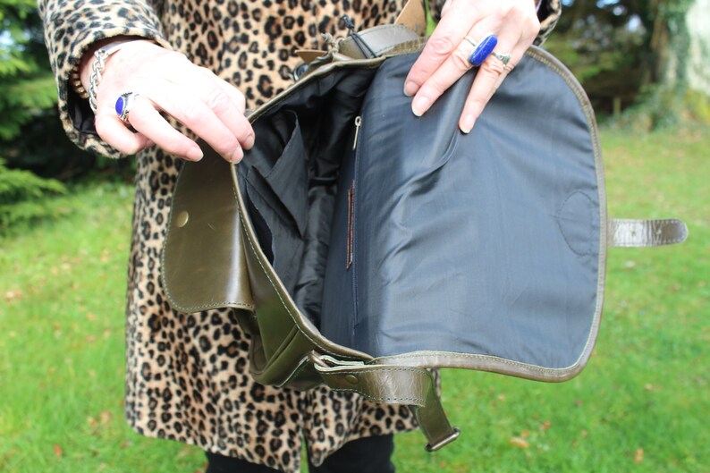 May include: A green leather satchel bag with a black fabric lining. The bag has a flap closure and a buckle strap. The bag is open and the lining is visible.