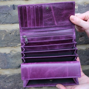 Puede incluir: Una cartera de cuero de color morado intenso con un diseño de acordeón de múltiples compartimentos. El interior incluye varias ranuras verticales para tarjetas, un bolsillo con cremallera y varias secciones abiertas, con cierre de botón.