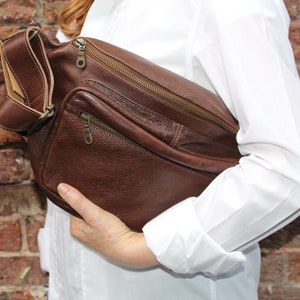 K&ouml;nnte beinhalten: Eine braune Leder-Umh&auml;ngetasche mit mehreren Rei&szlig;verschlussf&auml;chern. Die Tasche hat einen langen Riemen und wird &uuml;ber der Schulter getragen.