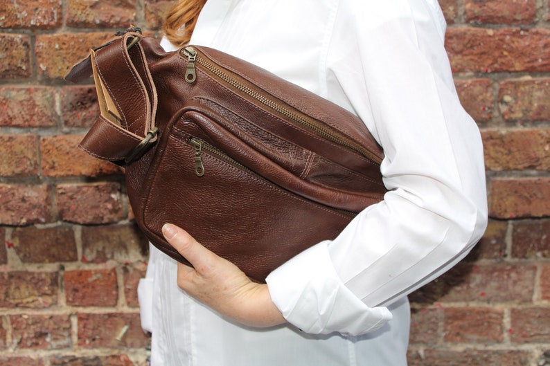 K&ouml;nnte beinhalten: Eine braune Leder-Umh&auml;ngetasche mit mehreren Rei&szlig;verschlussf&auml;chern. Die Tasche hat einen langen Riemen und wird &uuml;ber der Schulter getragen.