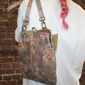 May include: A floral patterned leather purse with a brass clasp and a long strap. The purse is worn over the shoulder of a person wearing a white shirt.