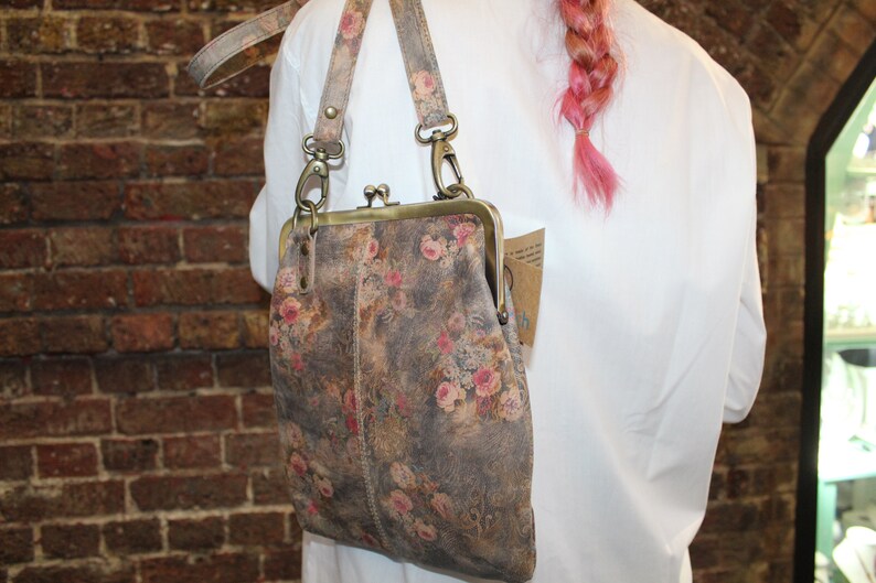 May include: A floral patterned leather purse with a brass clasp and a long strap. The purse is worn over the shoulder of a person wearing a white shirt.