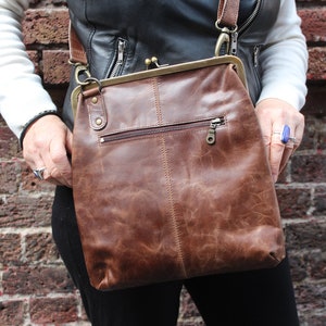 May include: A brown leather crossbody bag with a flap closure and a zippered pocket on the front. The bag has a distressed look and is worn over the shoulder.