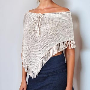 Puede incluir: Poncho blanco roto con hombros descubiertos y flecos. El poncho tiene un cierre de lazo en el cuello. La modelo lleva una falda vaquera azul oscuro. El fondo es una pared blanca lisa.