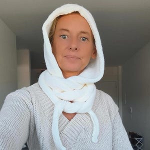 Könnte beinhalten: Ein weißes Strick-Set aus Kapuze und Schal. Die Kapuze ist hochgezogen und rahmt das Gesicht ein, der Schal ist um den Hals gewickelt. Das Set besteht aus einem dicken, strukturierten Strickstoff. Der Pullover ist hellbeige mit V-Ausschnitt.