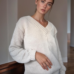 May include: A cream-coloured, knitted V-neck sweater. The sweater features a ribbed collar and cuffs, and a relaxed fit. The sleeves are long and the material appears to be a soft, chunky knit. The sweater is a neutral colour, suitable for various outfits.