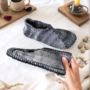 Peut inclure: Une paire de pantoufles en tricot gris et blanc avec une semelle en caoutchouc noir. Les pantoufles sont sur un couvre-lit blanc.