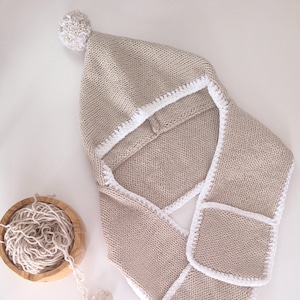 Könnte beinhalten: Beige Strickhaube mit weißer Einfassung und weißem Bommel. Die Haube hat eine schalförmige Verlängerung mit Taschen. Daneben eine Holzschale mit beiger Wolle. Ideal für kalte Tage.