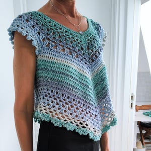 Könnte beinhalten: Handgefertigtes Häkeltop in Blau-, Türkis- und Weißtönen. Das Oberteil hat einen V-Ausschnitt und ein einzigartiges asymmetrisches Design. Die Ärmel haben einen gewellten Rand. Das offene Webmuster ist im gesamten Kleidungsstück sichtbar.