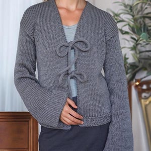 Puede incluir: Un cárdigan de punto gris con cuello en V y cierre de lazo. El suéter de manga larga tiene una textura acanalada y se usa sobre una camiseta azul claro. La modelo está de pie frente a una cortina blanca y una puerta de madera.