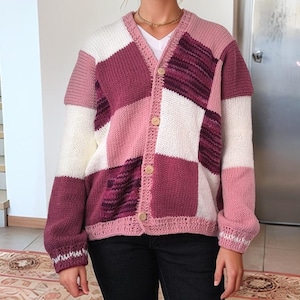 Può includere: Cardigan patchwork nei toni del rosa, bianco e bordeaux. Il maglione presenta uno scollo a V, maniche lunghe e bottoni in legno. Il design è un mix di quadrati a maglia lisci e strutturati.