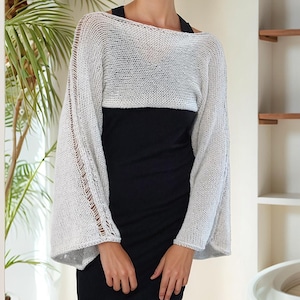 May include: A light gray, open-weave bolero sweater with wide sleeves. The sweater is worn over a black dress. The sleeves have a decorative open stitch detail. The background includes a palm plant and a white wall.