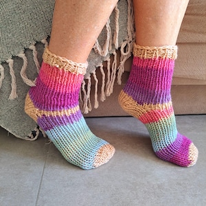 Op de afbeelding: Een paar kleurrijke gestreepte gebreide sokken met een beige boord. De sokken zijn gemaakt van verschillende kleuren, waaronder roze, paars, blauw en groen.