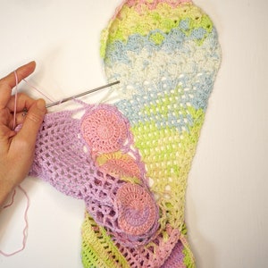 May include: A person is crocheting a colorful, multi-colored crochet blanket with a pink, green, and yellow color scheme. The blanket is made with a variety of stitches, including a crochet shell stitch and a crochet granny square stitch.